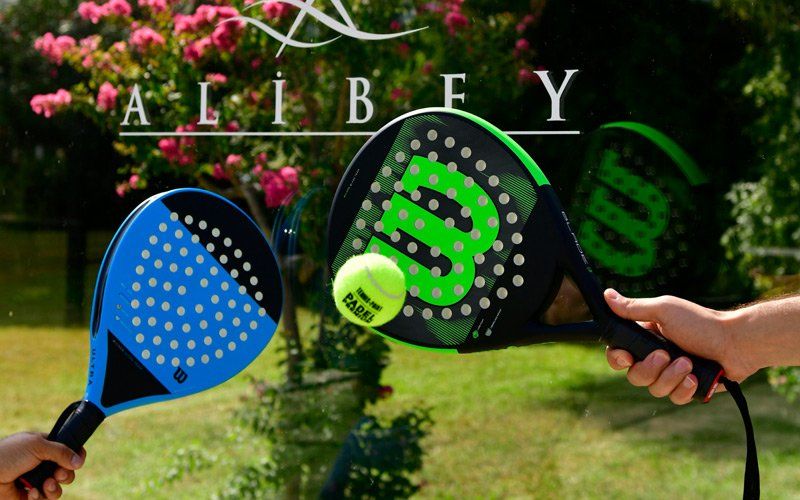 Zwei Padel Tennisschläger mit Ball vor Ali Bey Logo auf Glaswand